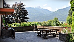 A patio with wooden benches and a stone floor, overlooking a mountainous landscape with dense greenery. - Enzianbrenner Bed & Breakfast (Hermagor-Pressegger See)