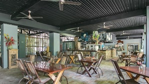 A spacious restaurant with high ceilings, exposed beams, and multiple ceiling fans. Tables and chairs are arranged in a casual layout, with a brick wall and a large window in the background. - Daintree Wild Zoo & B&B (Lower Daintree)