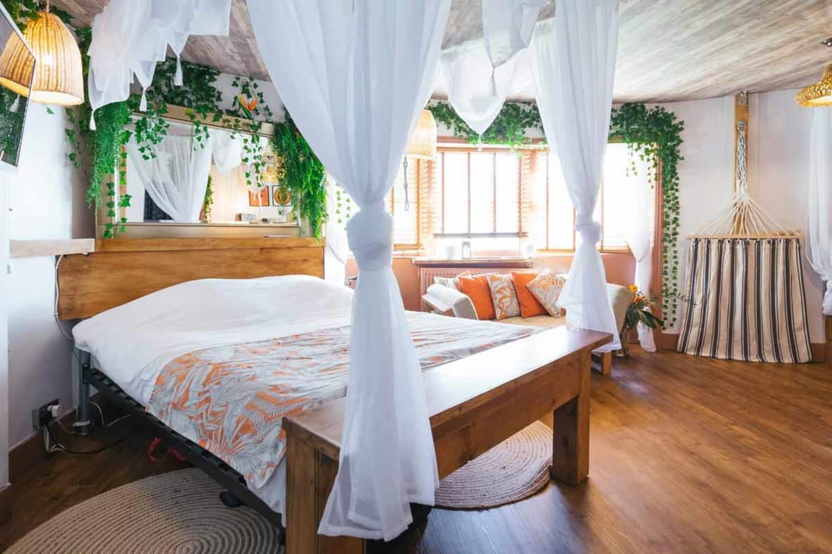 Ein Schlafzimmer mit einem hölzernen Bett, einem weißen Baldachin und Blick auf eine Wohnbereich mit einer Couch und gestreiften Vorhängen.