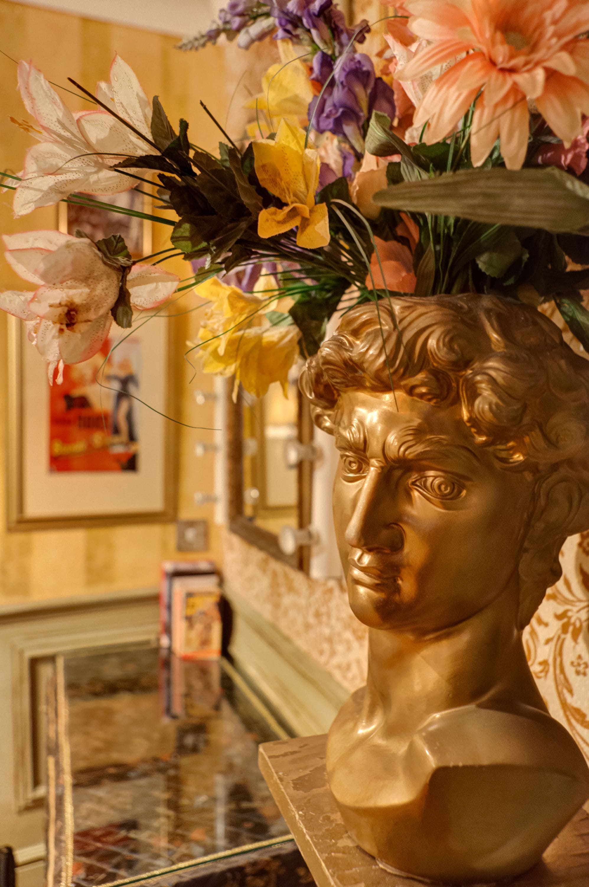 A marble bust with a floral arrangement on top, set against a wall with framed pictures and a marble countertop.