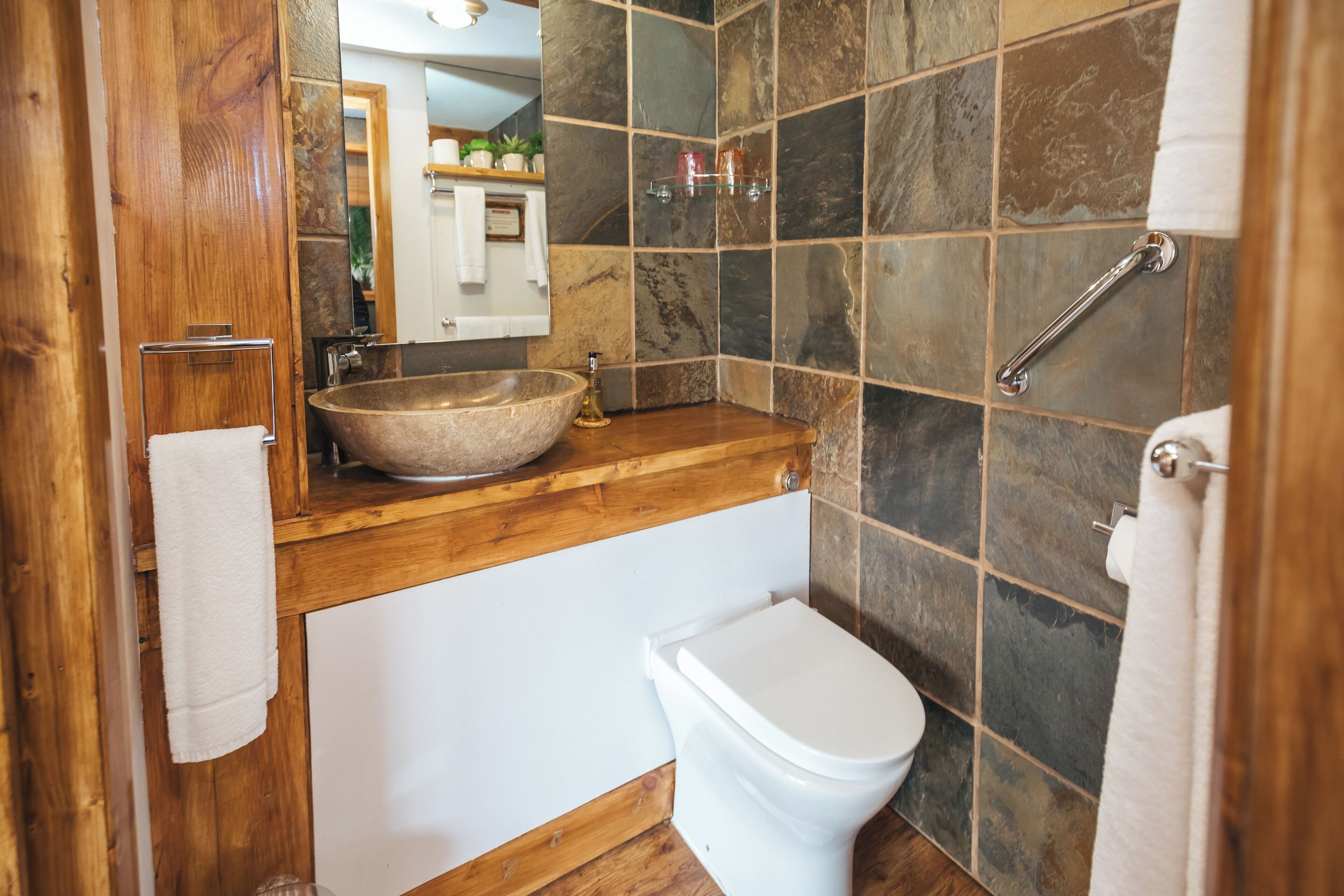 A bathroom with a wooden vanity, a stone sink, a white toilet, and a glass shelf with toiletries.