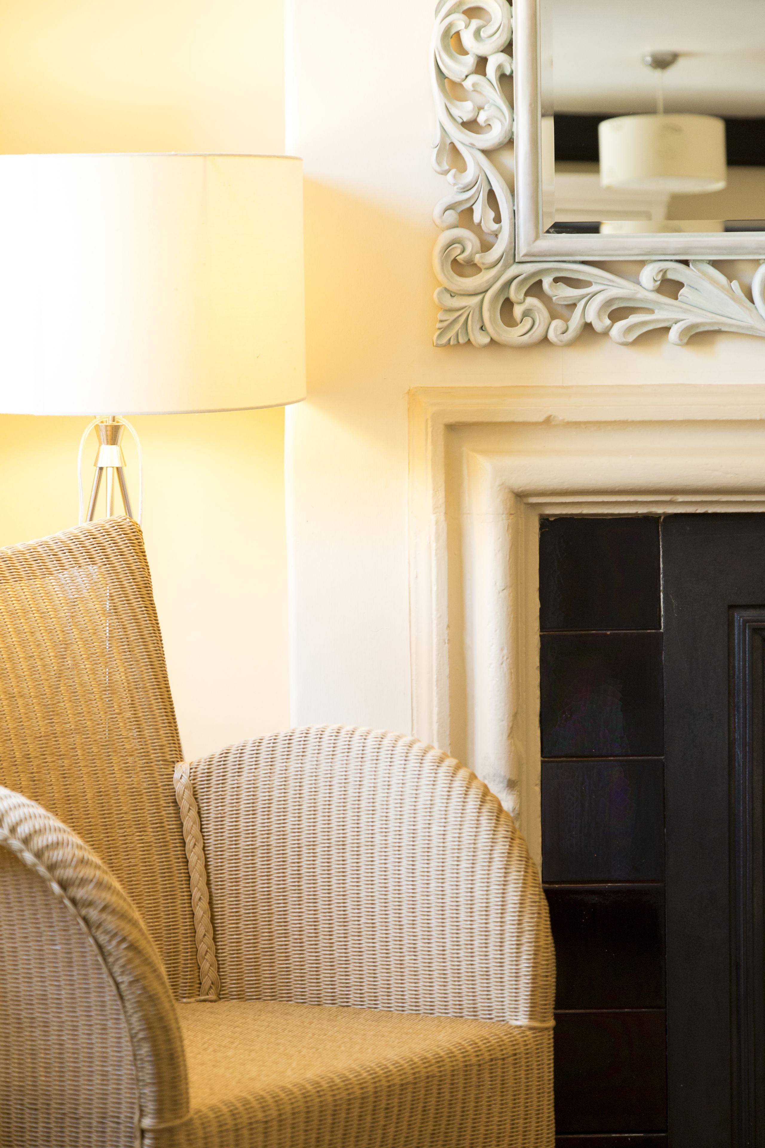 A wicker chair with a woven texture, positioned next to a wall with a white ornate mirror and a black fireplace.
