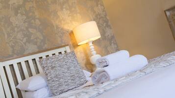 A neatly made bed with pillows and rolled towels, a wall with floral wallpaper, and a bedside lamp.