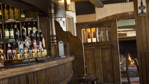 A bar area with wooden bar counter and stools, shelves with various bottles, and a fireplace in the background. - Red Lion - B&B (Chippenham)
