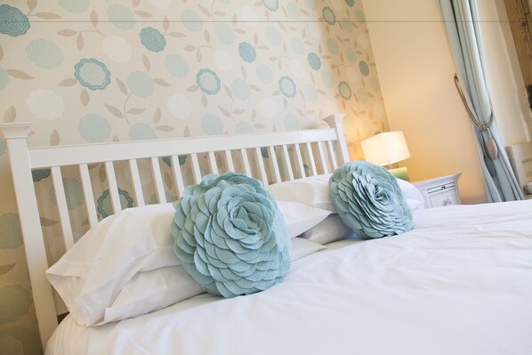 Una cama bien hecha con ropa de cama blanca y almohadas decorativas azules, un cabecero blanco y una mesita de noche con lámpara.