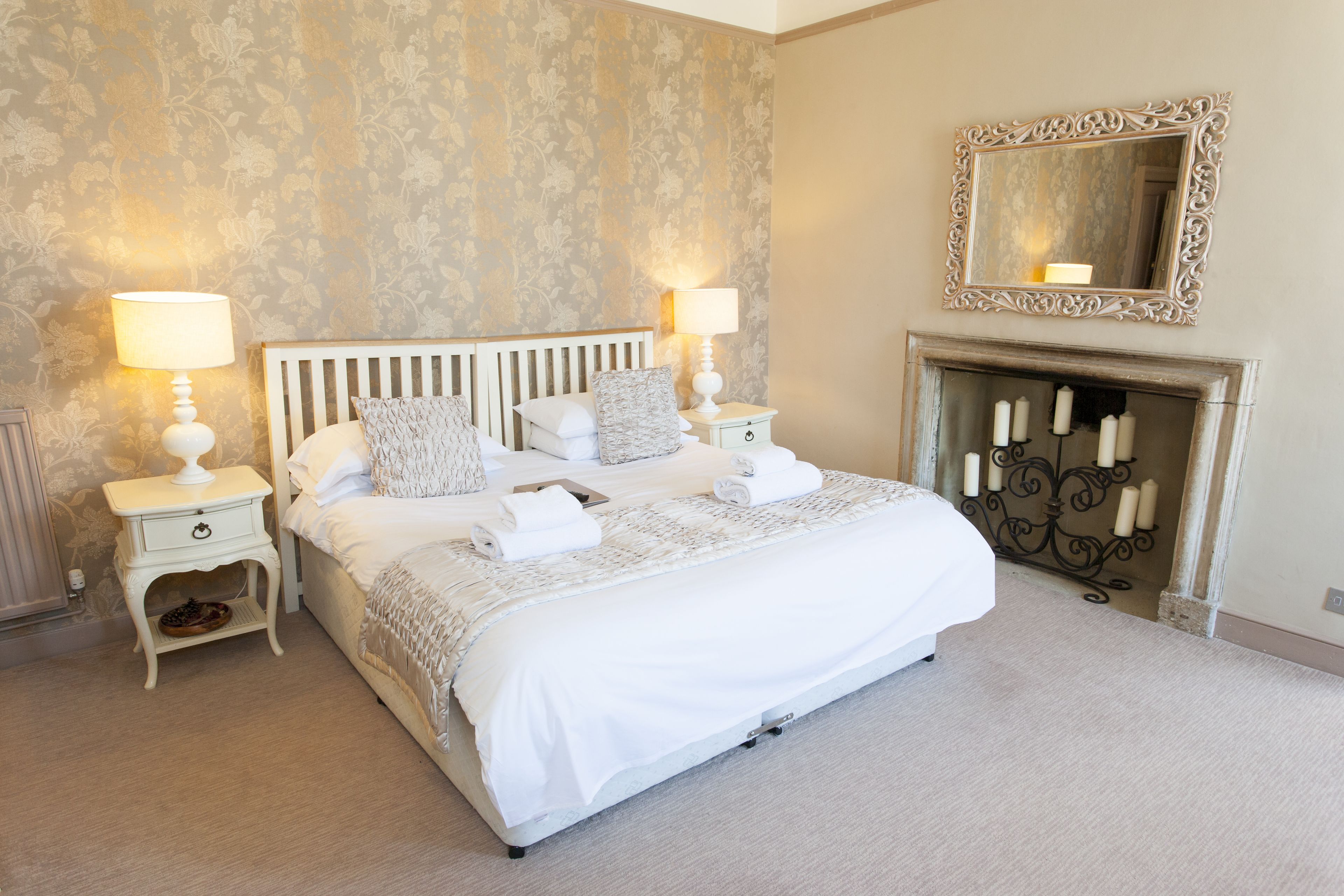 A neatly made bed with white linens, two bedside tables with lamps, a fireplace with candles, and a mirror on the wall.
