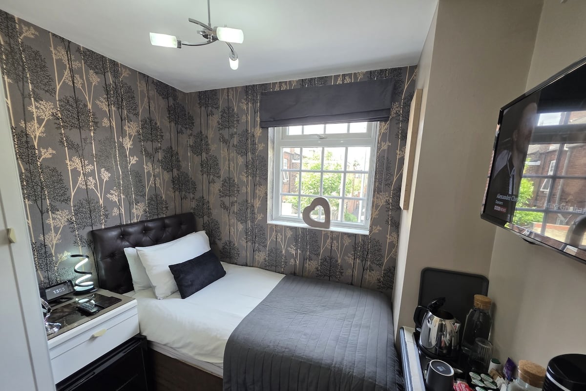 A bedroom with a bed, a nightstand, a TV, a window with a view, and a desk with a lamp.