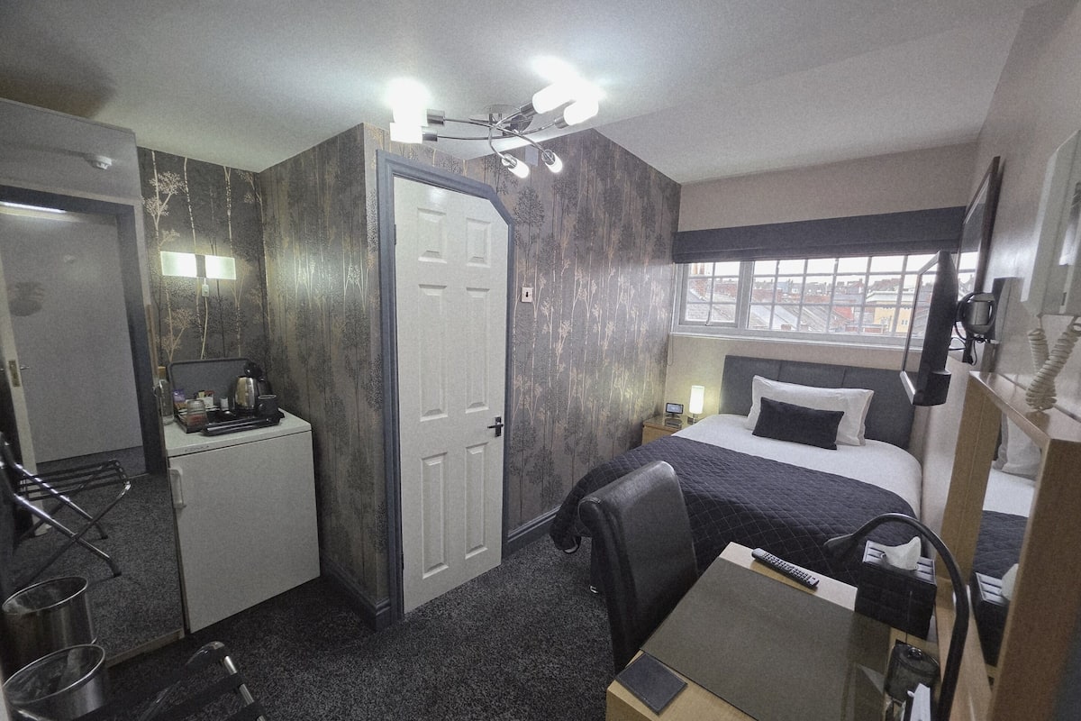 Een kleine hotelkamer met een bed, bureau, stoel en een klein kitchenette.