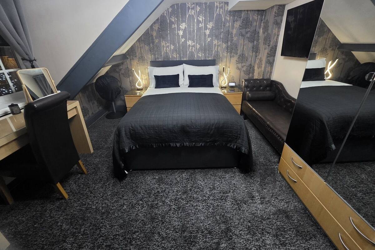 Een moderne slaapkamer met een groot bed, een bureau en een stoel. Er is een spiegel, een nachtkastje en een lamp.
