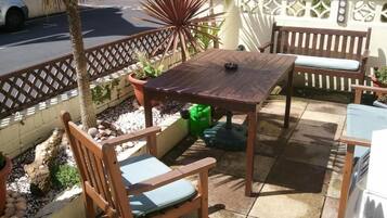 Un patio al aire libre con una mesa y sillas de madera, rodeado de plantas en macetas y una valla decorativa.