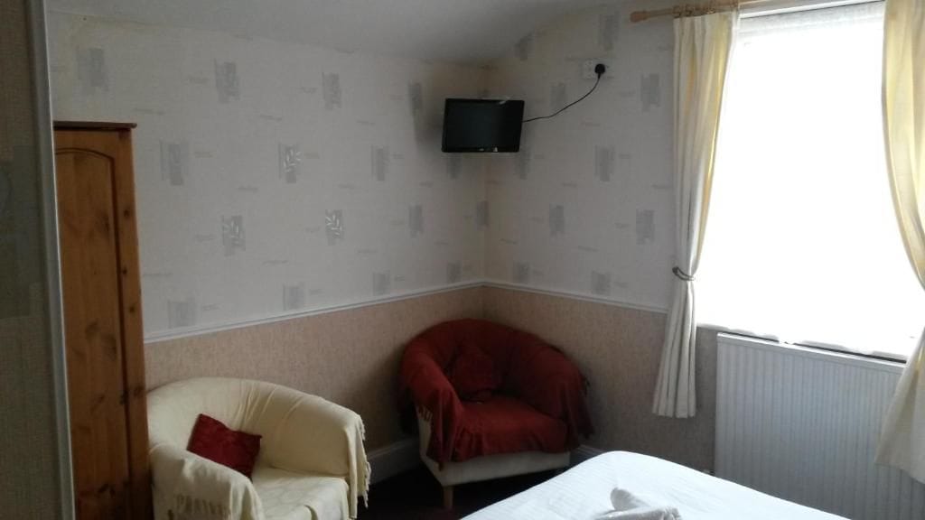 A room with a bed, a red chair, a white armchair, a television mounted on the wall, and a window with curtains.