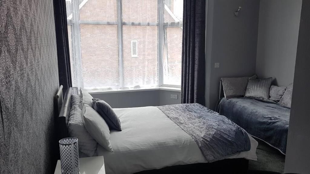 A bedroom with a large window, a bed with a blue blanket, and a bedside table with a lamp.