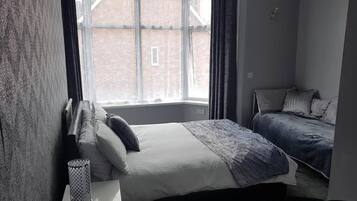 A bedroom with a large window, a bed with a blue blanket, and a bedside table with a lamp.
