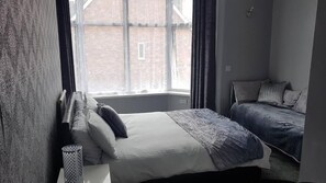 A bedroom with a large window, a bed with a blue blanket, and a bedside table with a lamp. - Brampton Guest House (Paignton)