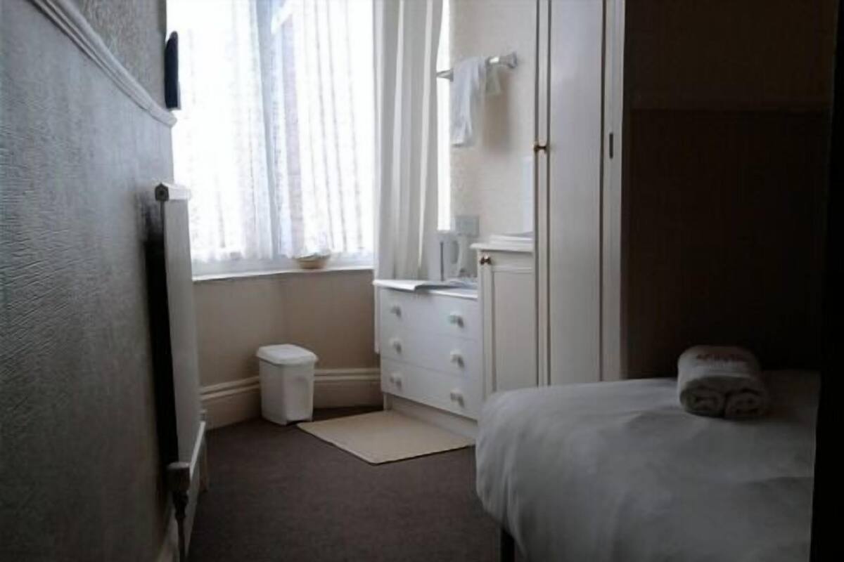 Une petite chambre propre, avec un lit simple, une table de chevet, une petite fenêtre avec des rideaux, une commode blanche et une poubelle blanche.