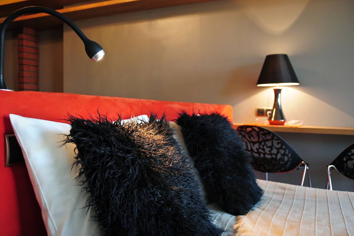 A bed with a red headboard, black furry pillows, and a white pillow. There is a black lamp on the bedside table and a red lamp on a shelf.