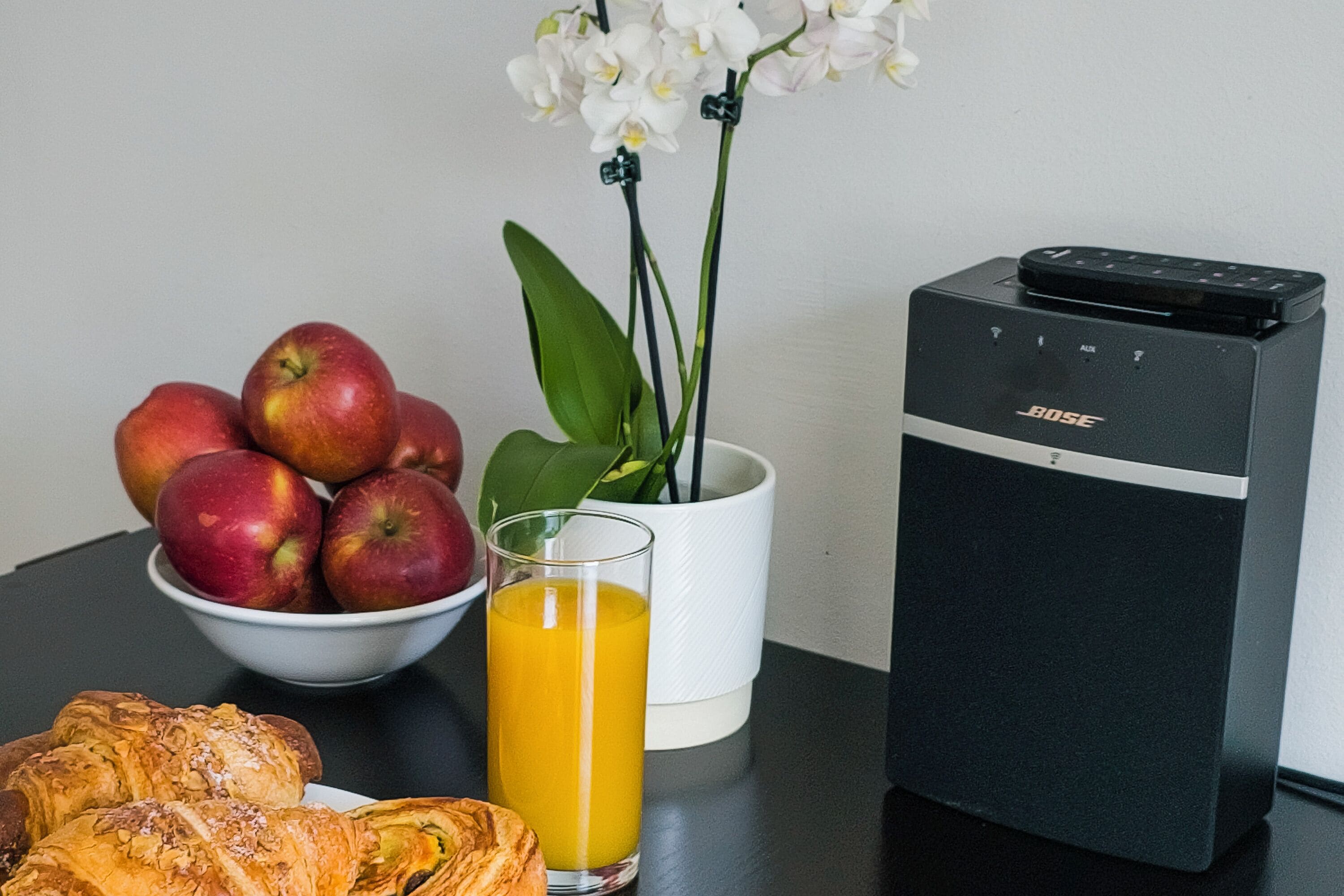 Una mesa con un bol de manzanas, un vaso de jugo de naranja, una orquídea en maceta y un altavoz.