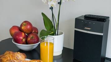 Una mesa con un bol de manzanas, un vaso de jugo de naranja, una orquídea en maceta y un altavoz.