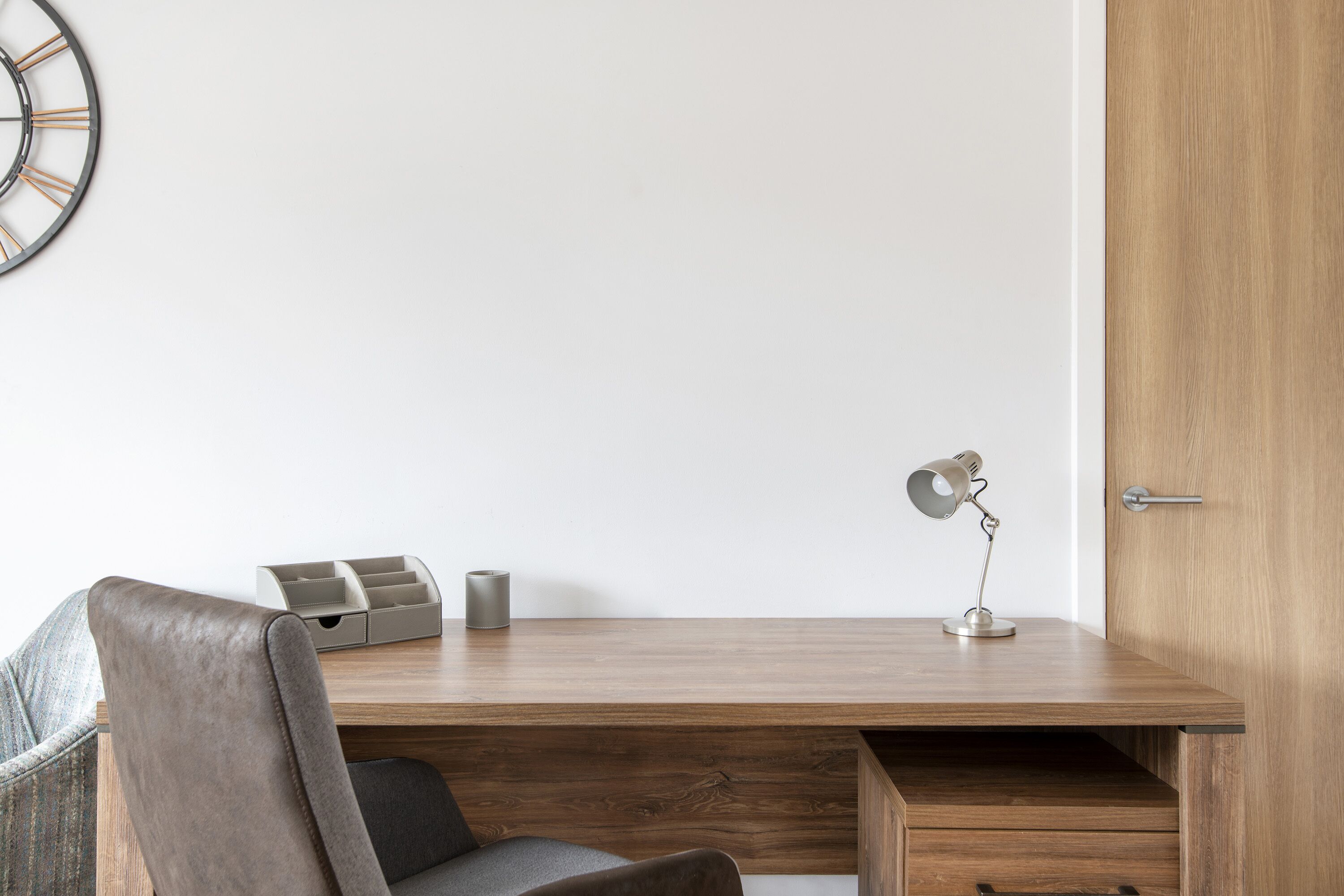 A modern office desk with a wooden finish, a chair, a desk lamp, and a wall clock.