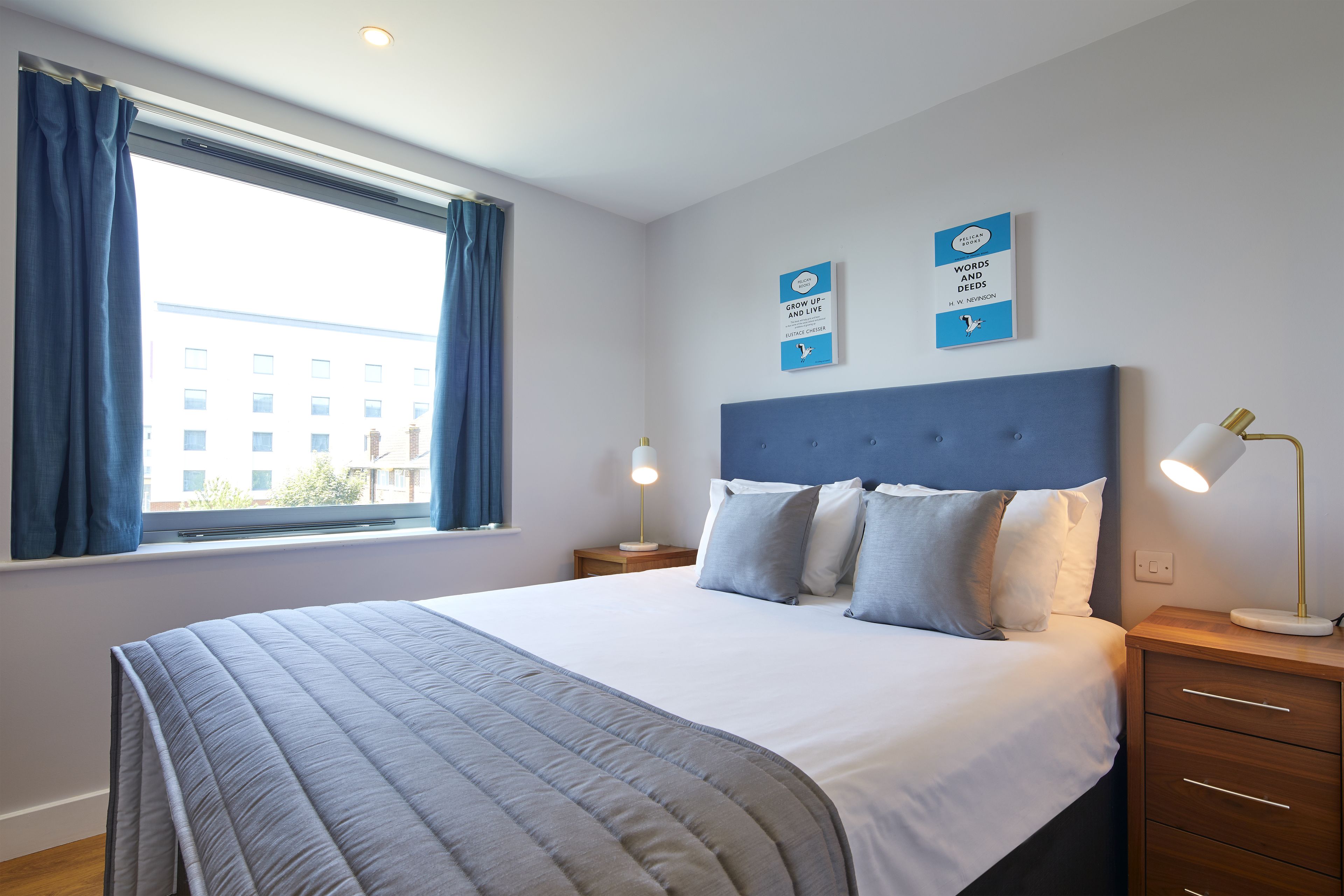 A neatly made bed with a blue headboard, a bedside lamp, and a window with curtains.
