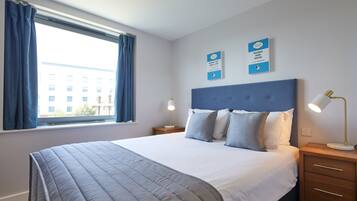 A neatly made bed with a blue headboard, a bedside lamp, and a window with curtains.