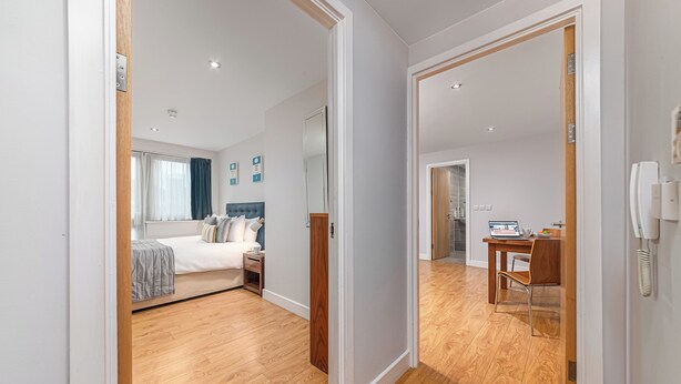 A modern hotel room with a wooden floor, a bed with a grey blanket, a bedside table, and a desk with a laptop. - HQ Aparthotel Farnborough (Farnborough)