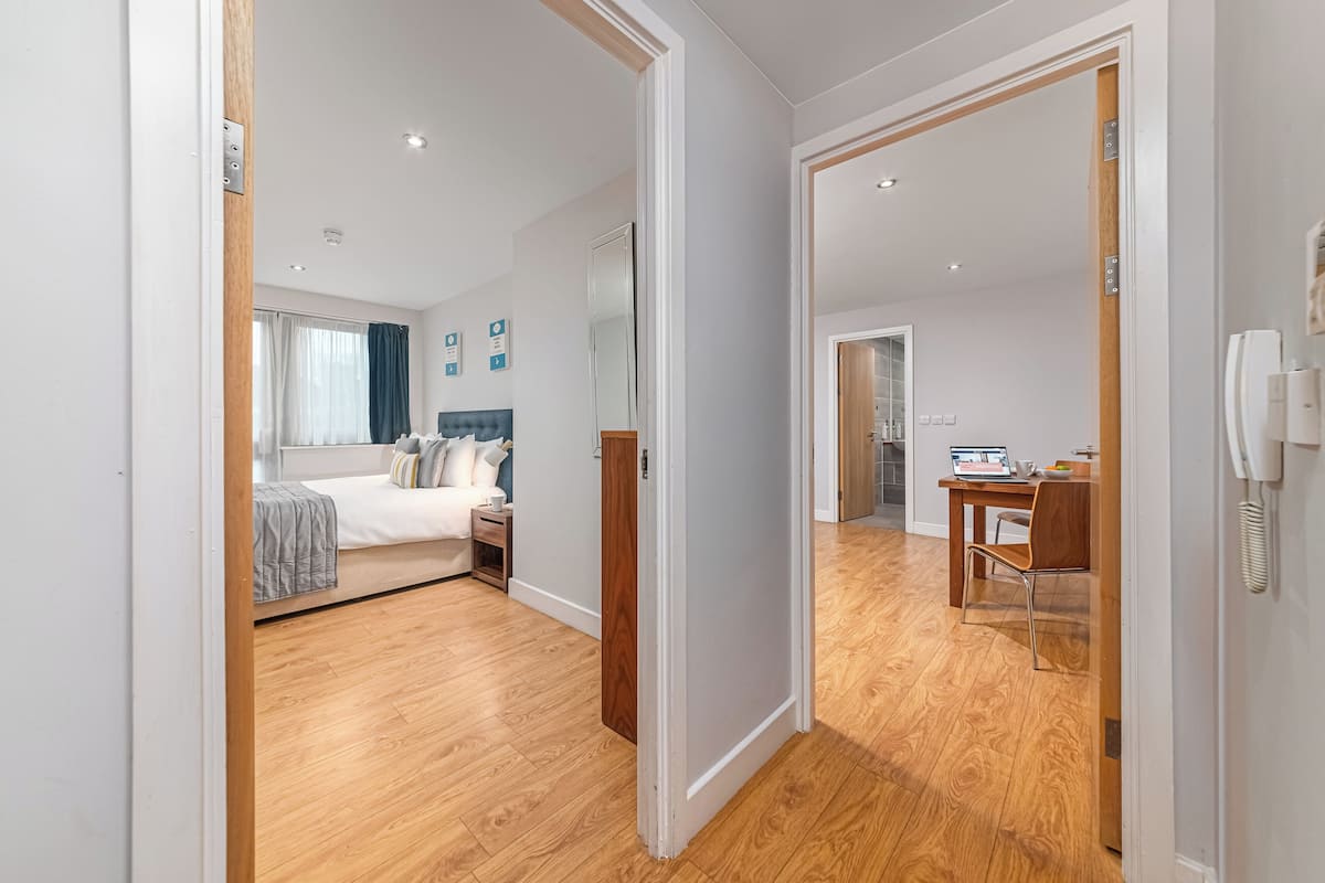 Une chambre d’hôtel moderne avec un plancher en bois, un lit recouvert d’une couverture grise, une table de chevet et un bureau avec un ordinateur portable.