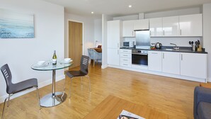 A modern kitchen with white cabinets, a stainless steel refrigerator, and a microwave. There is a dining area with a glass table and two chairs. - HQ Aparthotel Farnborough (Farnborough)