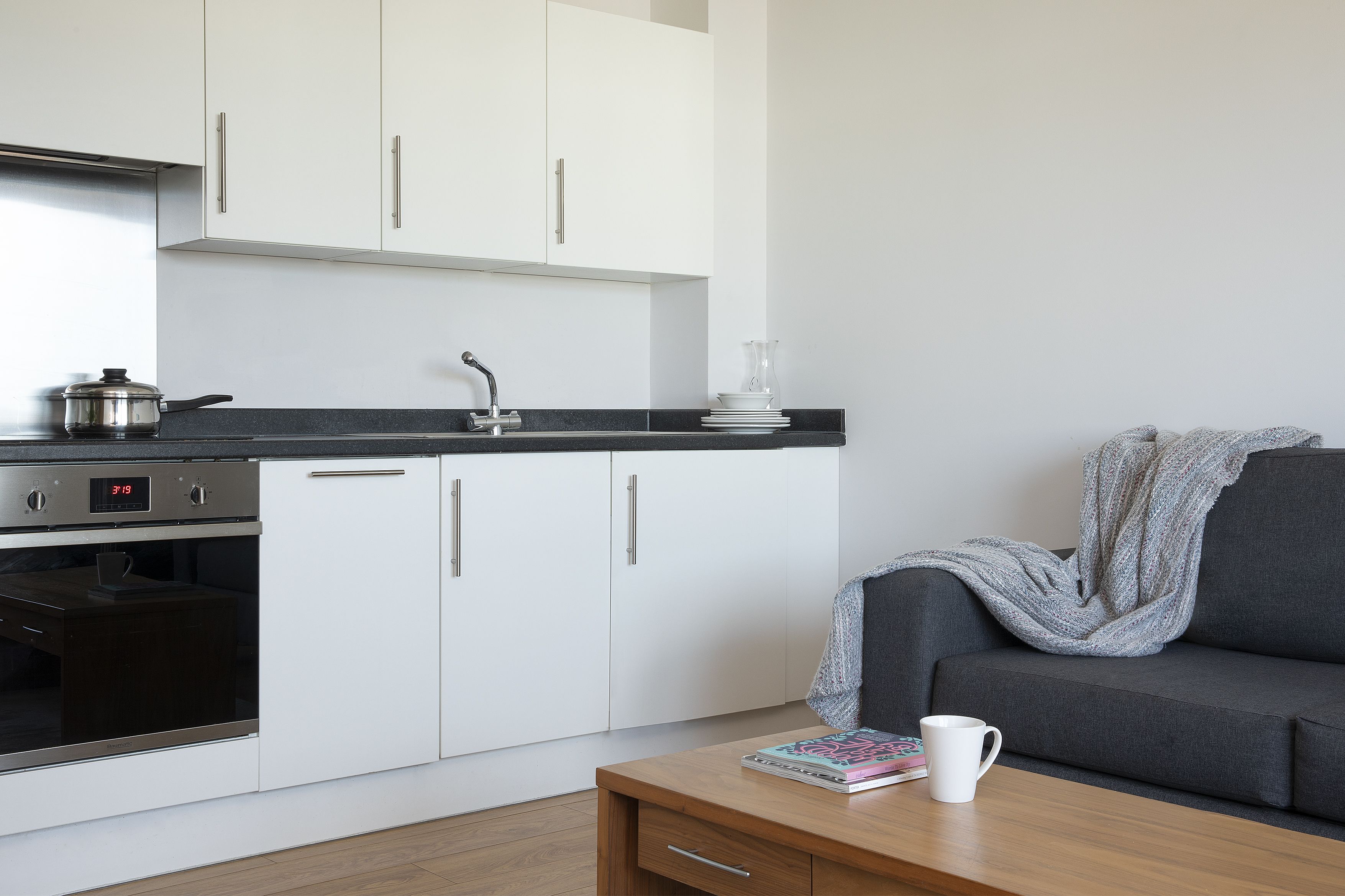 A modern kitchen with white cabinets, a stove, and a sink. There's a grey sofa with a blanket and a white mug on a wooden table.