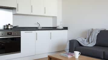 A modern kitchen with white cabinets, a stove, and a sink. There's a grey sofa with a blanket and a white mug on a wooden table.