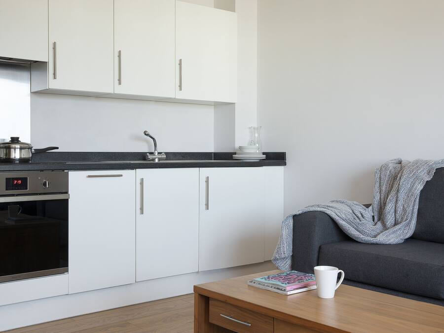 A modern kitchen with white cabinets, a stove, and a sink. There's a grey sofa with a blanket and a white mug on a wooden table.