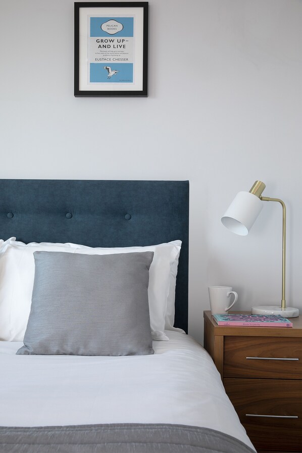 A neatly made bed with a blue headboard, a framed book on the nightstand, and a white lamp. - HQ Aparthotel Farnborough (Farnborough)
