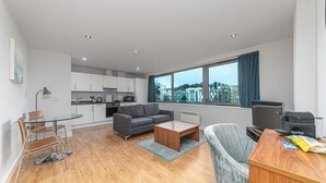 A modern apartment with a kitchen, living room, and dining area. It features a large window with a city view, a gray sofa, a wooden coffee table, and a dining table with chairs. - HQ Aparthotel Farnborough (Farnborough)