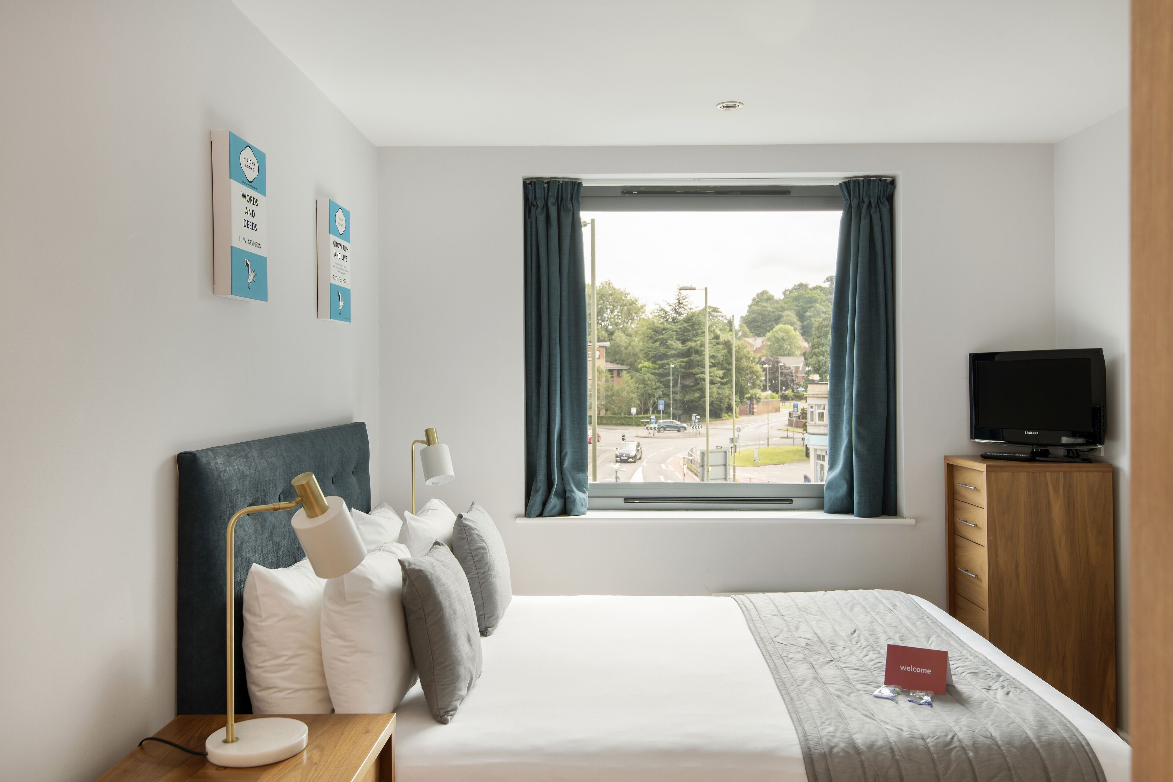 A hotel room with a bed, a desk with a lamp, a television, and a window with curtains.