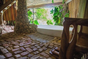 Un piso de baldosas de piedra, un pilar de piedra, una silla de madera y una ventana con vistas al verdor.