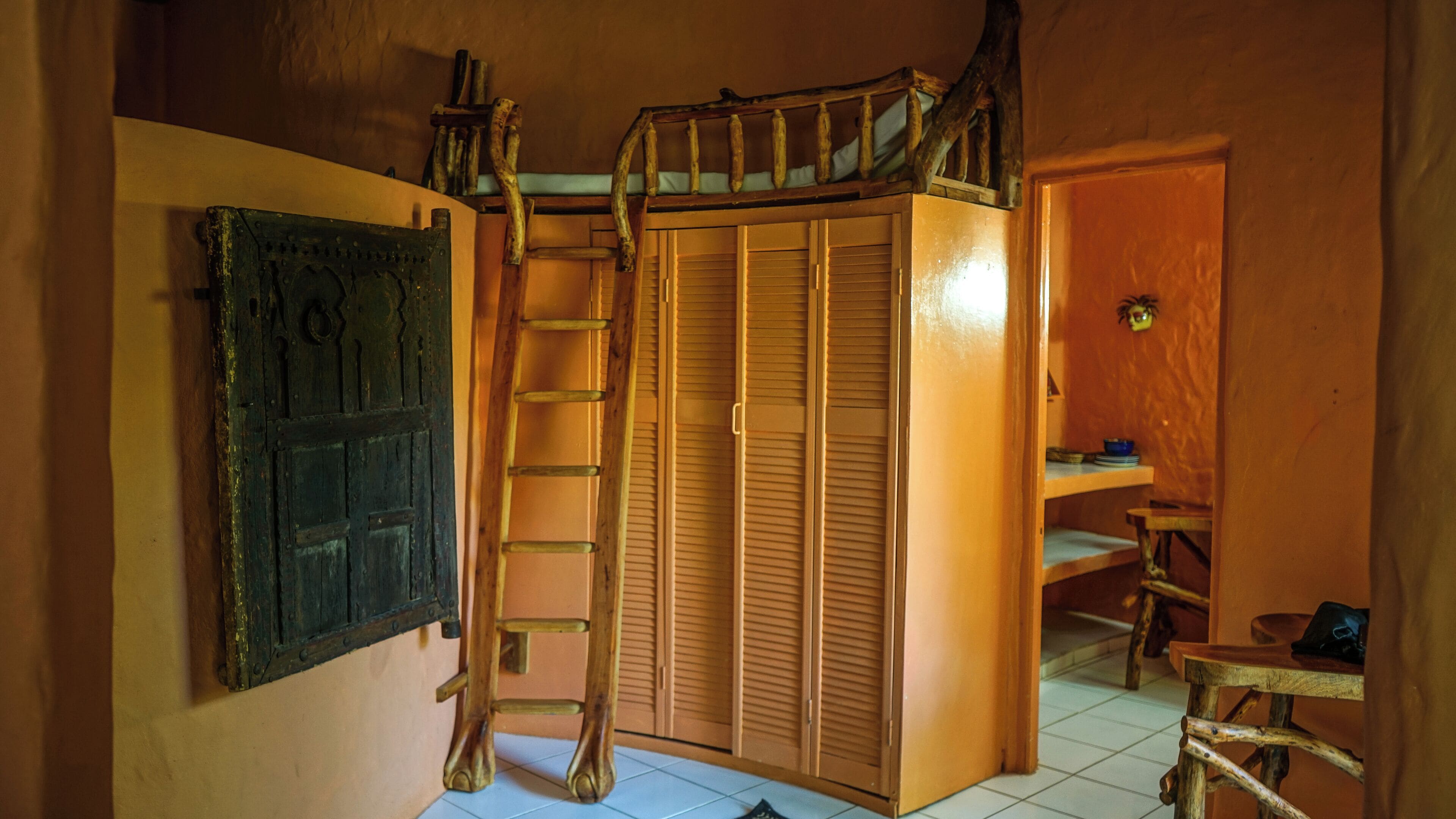 A room with a wooden ladder leading to a lofted sleeping area, a wardrobe, and a small table with a chair.