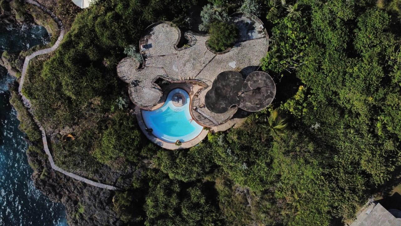 An aerial view of a resort with a circular pool surrounded by lush greenery and rocky terrain.