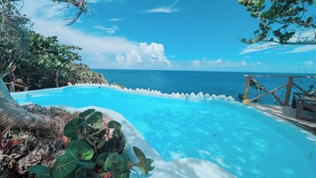 Piscina azul cristalina con vista al océano, rodeada de vegetación tropical y una terraza de madera.