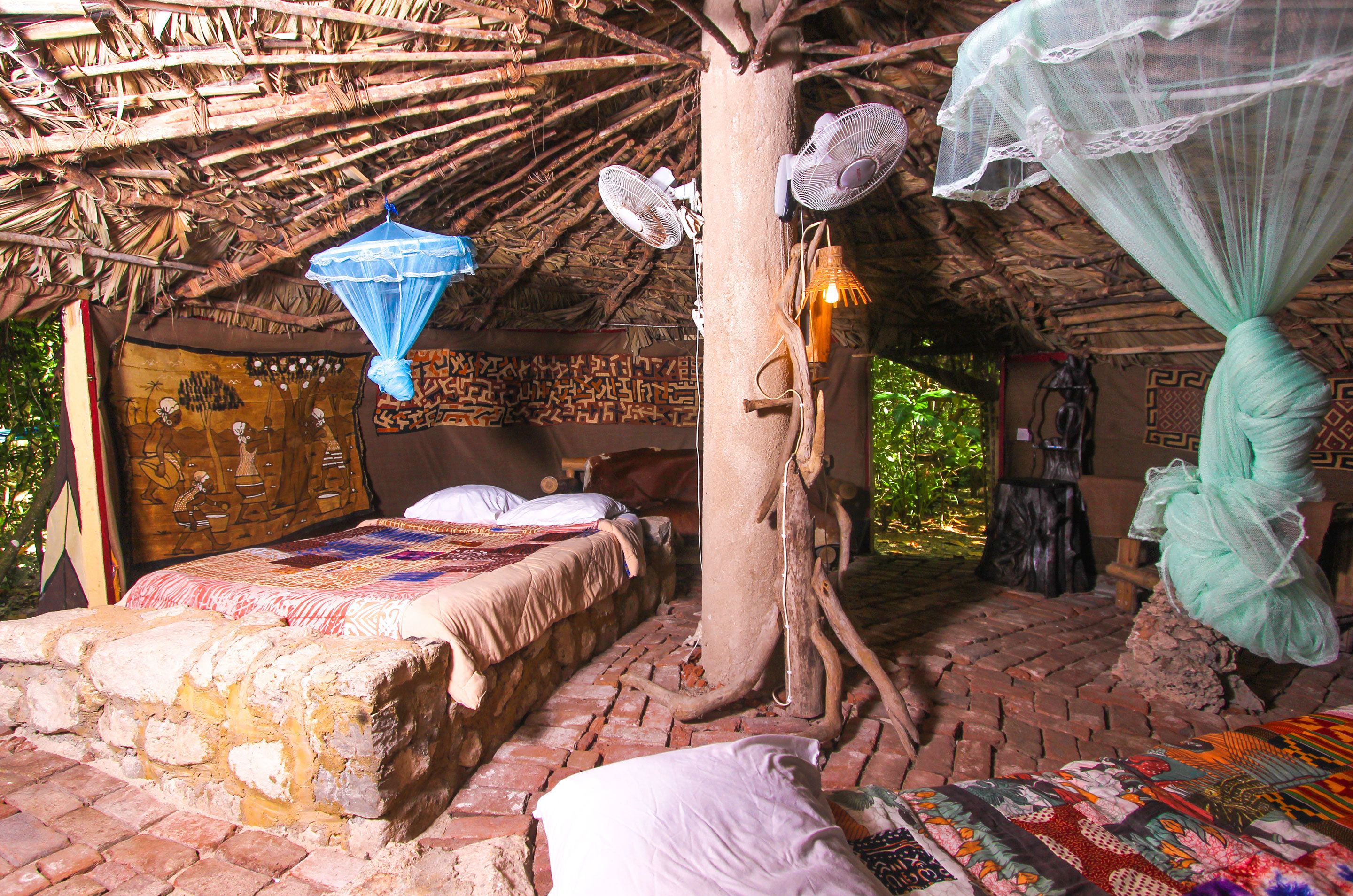 Une pièce rustique avec un toit de chaume, un lit recouvert d’une couverture aux couleurs vives, un ventilateur suspendu et un tronc d’arbre servant de support à un baldaquin.