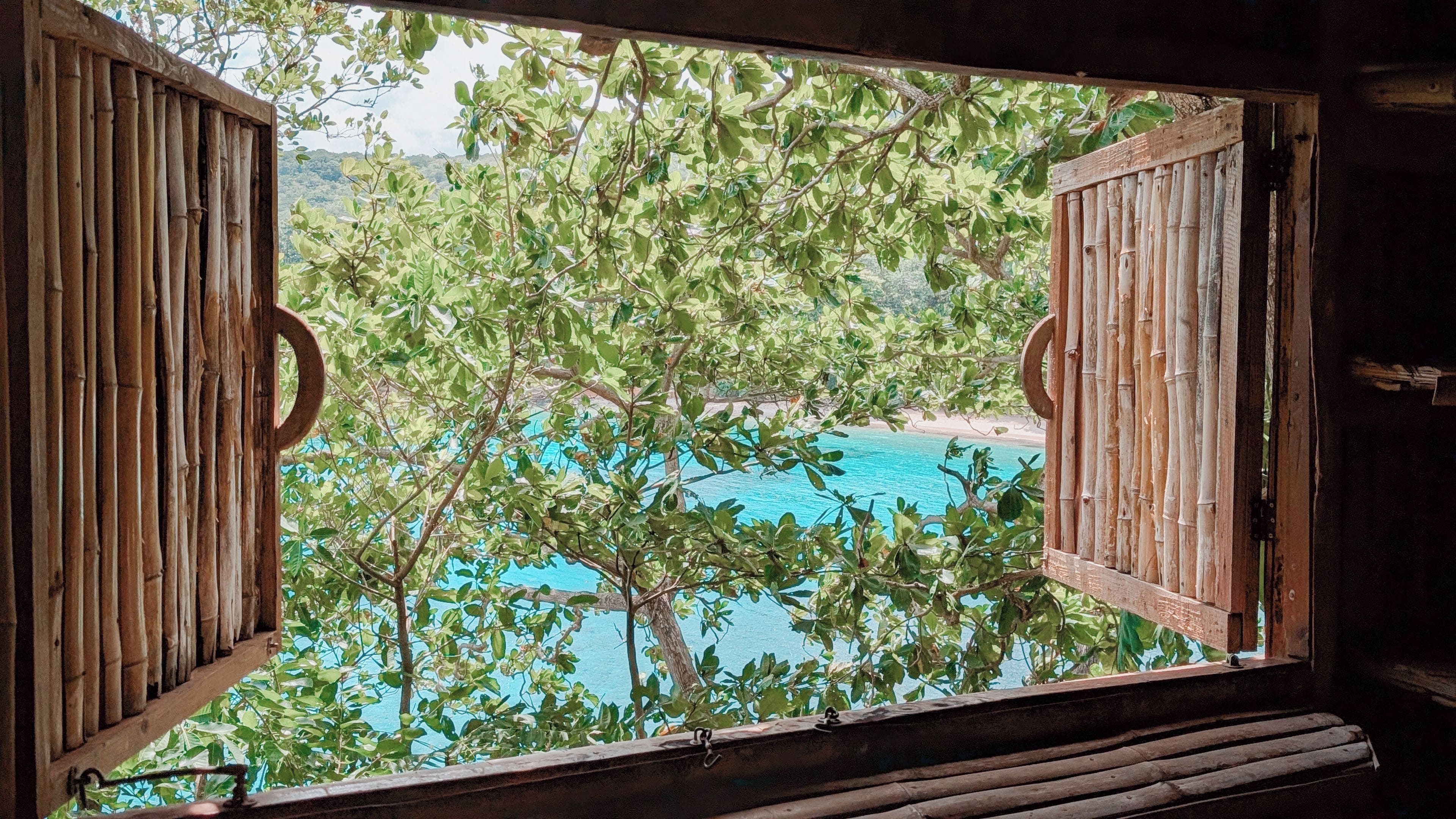 A wooden-framed window with bamboo slats, overlooking a beach and clear blue water.