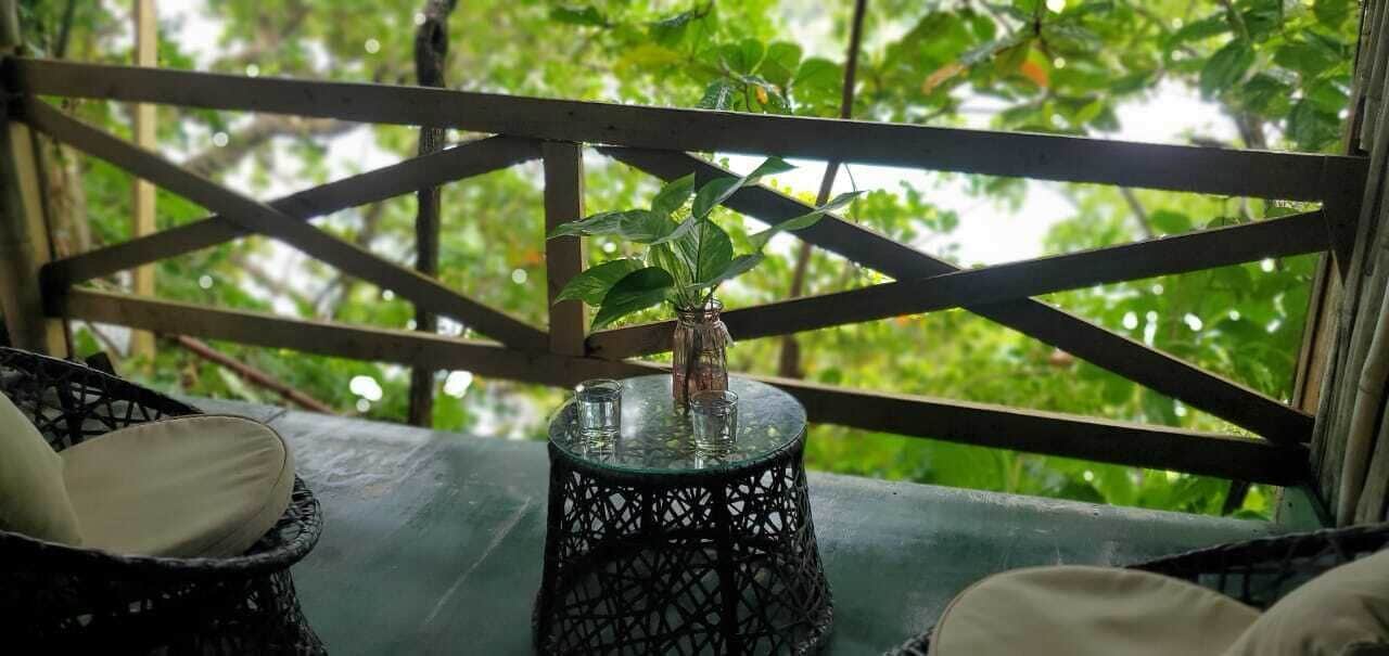 A wooden deck with a glass table and chairs, surrounded by lush greenery.