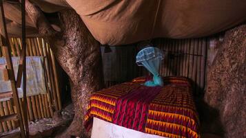 A rustic room with a bed under a large tree, bamboo walls, and a simple mosquito net.