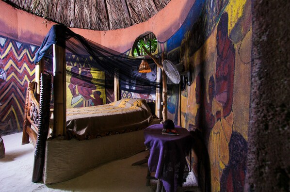 A room with a bed, a small table, and a fan. The walls are decorated with colorful murals.