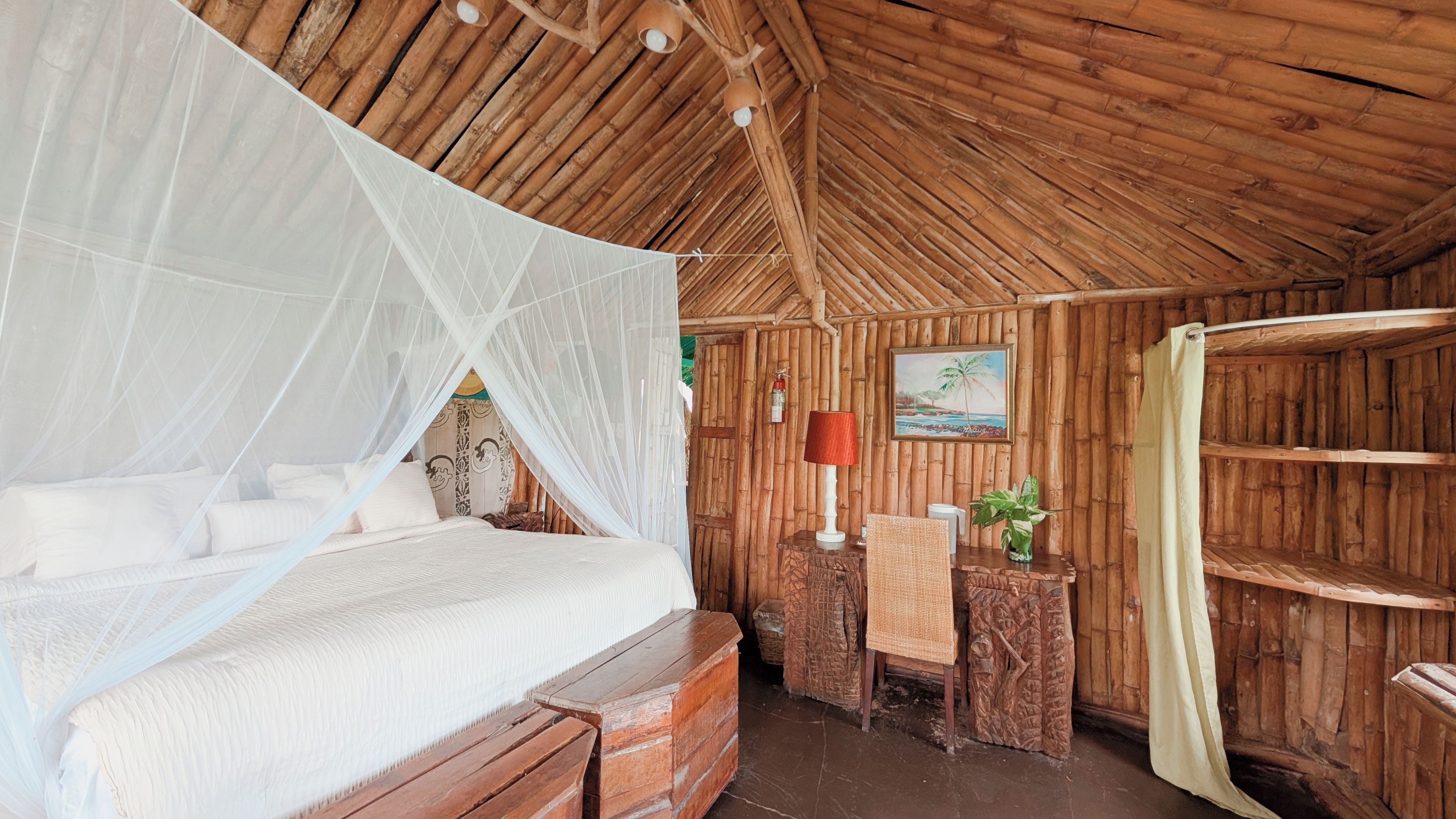 A room with a bed under a mosquito net, wooden walls, a painting, and a small table with a lamp.