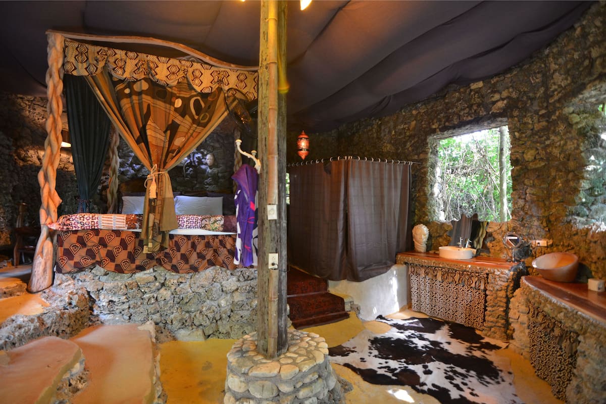 A room with a stone wall, a stone floor, a bed with a canopy, a wooden table, a window with curtains, and a cowhide rug.