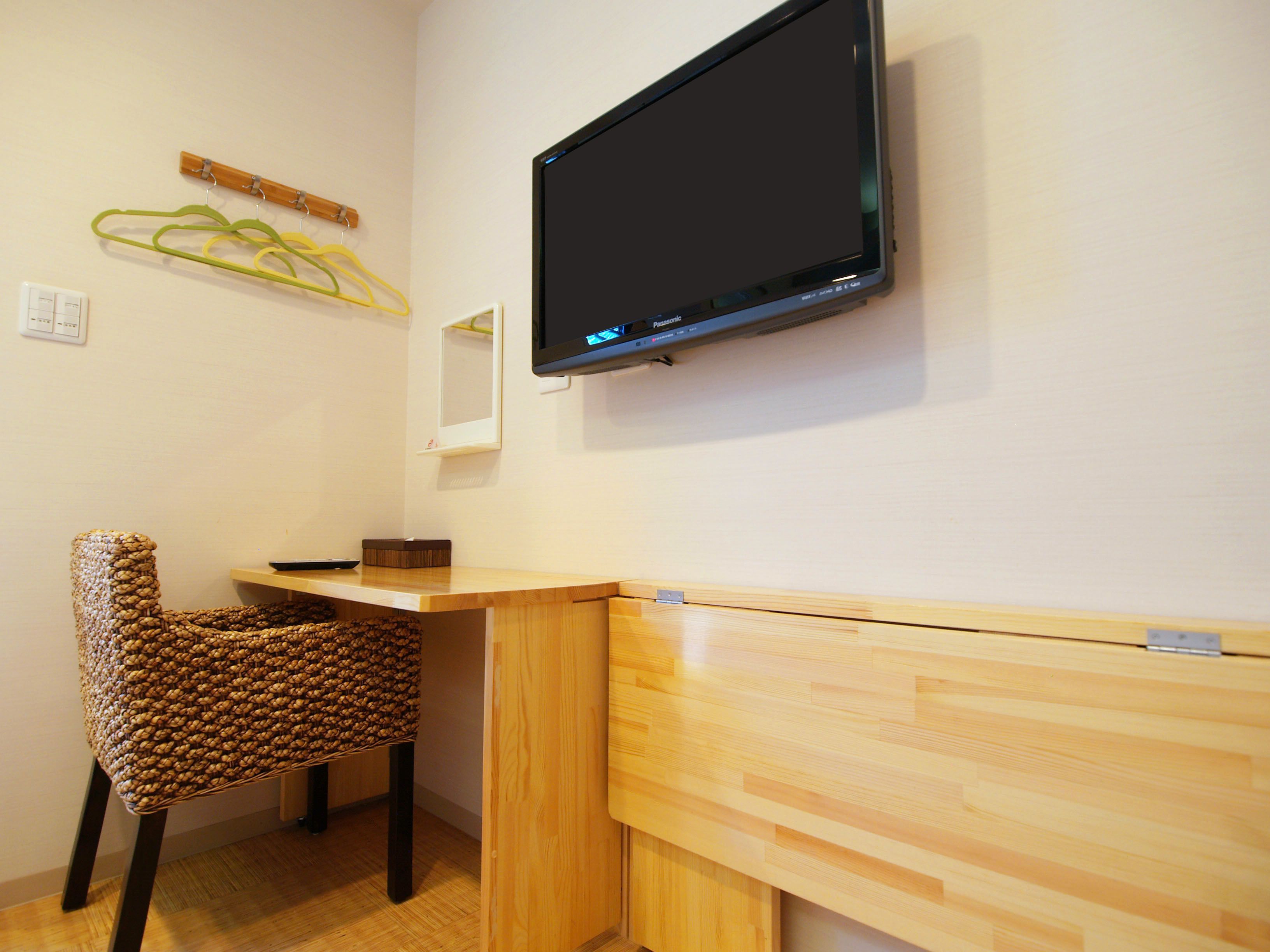 A room with a wooden desk, a woven chair, a mounted television, and a wall-mounted mirror.