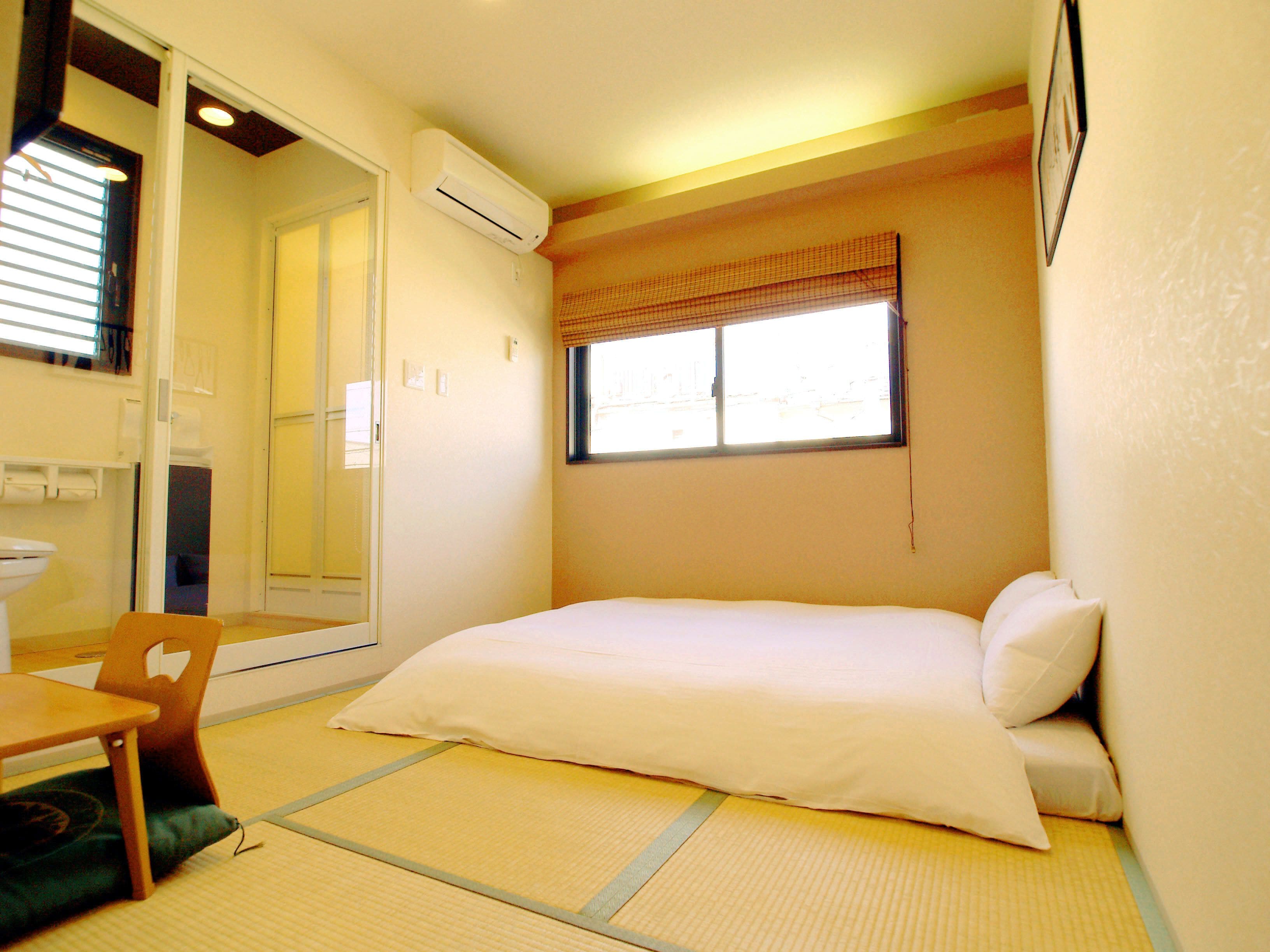 A small, compact room with a bed, a chair, a toilet, and an air conditioning unit.