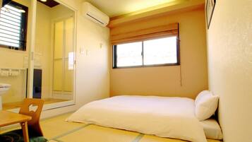 A small, compact room with a bed, a chair, a toilet, and an air conditioning unit.