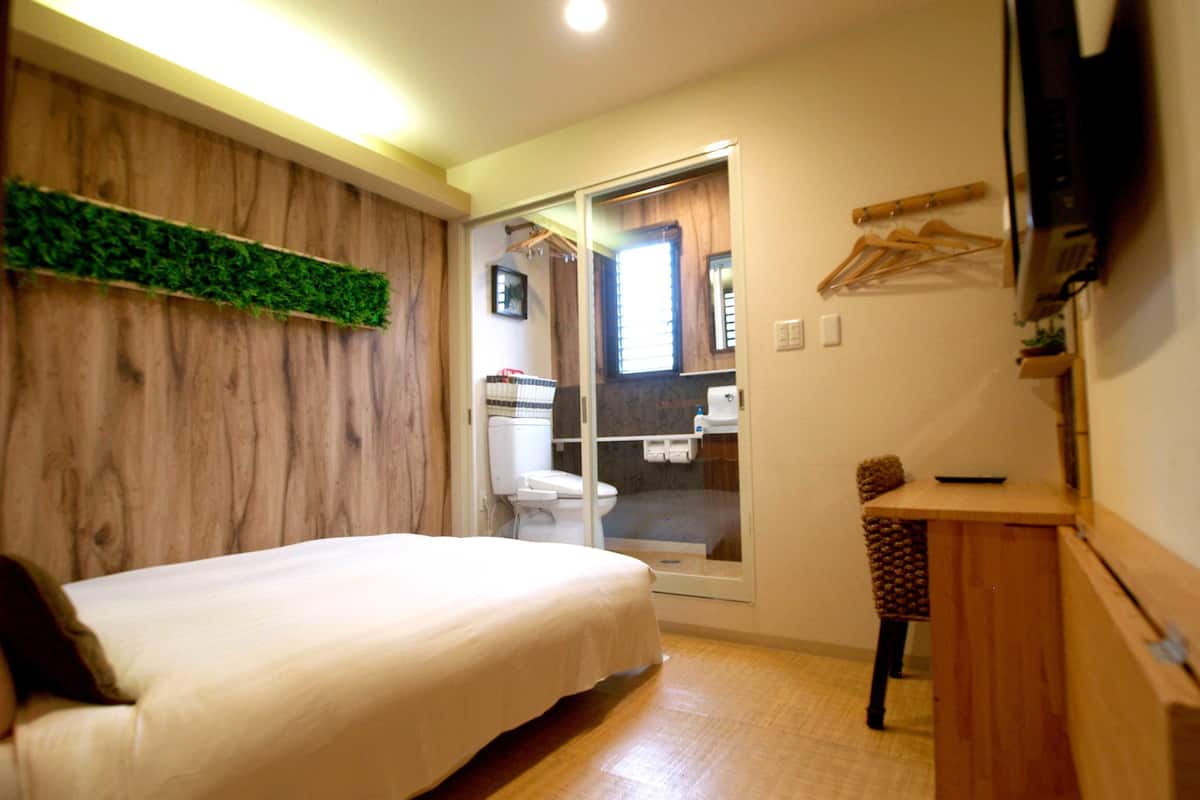 A bedroom with a bed, a wooden wardrobe, a small desk, and a television on the wall.