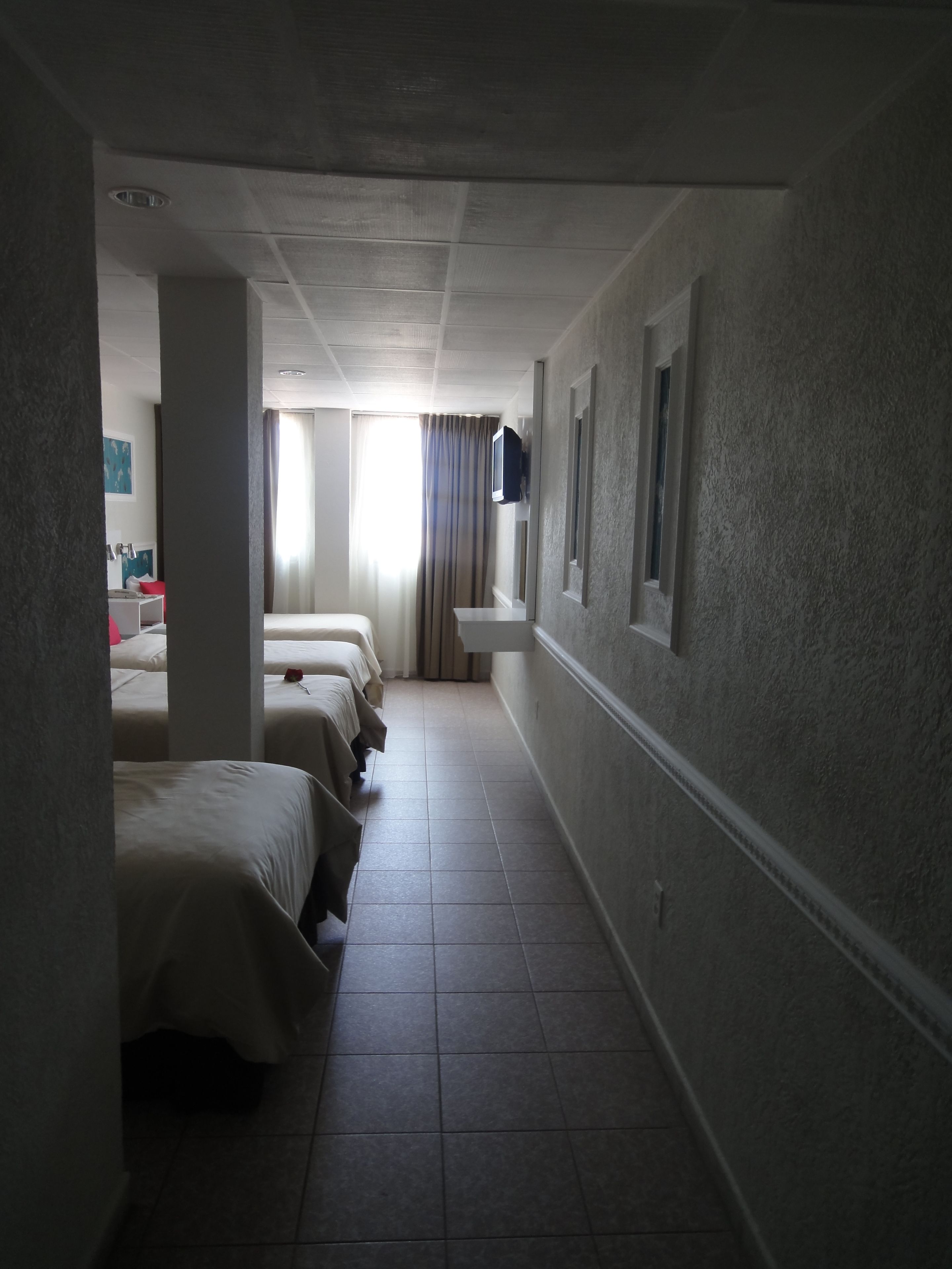 Et hotelværelse med tre senge langs væggen, et lille håndvaskområde og et fjernsyn monteret på væggen.